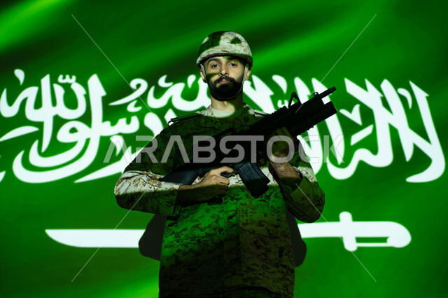مفهوم حب الوطن والانتماء إليه، الوقوف بثقة والنظر إلى الكاميرا، انعكاس ضوء علم السعودية على الجسم، إحياء ذكرى اليوم الوطني السعودي 23 سبتمبر، الاحتفال بالعيد الوطني، يوم العلم 11 مارس، جندي عربي خليجي يرتدي الزي العسكري والخوذة يحمل السلاح.