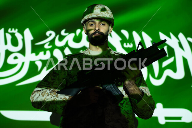 Standing confidently and looking at the camera, the light of the flag reflected on the face, commemorating the Saudi National Day, September 23, celebrating the National Day, Flag Day, March 11, a close-up of a Gulf Arab soldier wearing a military uniform and helmet carrying a weapon, the concept of love for the homeland and belonging to it.