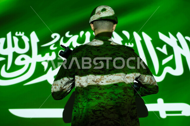 إحياء ذكرى اليوم الوطني السعودي 23 سبتمبر، انغماس الجسم في ضوء علم المملكة العربية السعودية، الاحتفال بالعيد الوطني ، يوم العلم 11 مارس، صورة من الخلف لجندي عربي خليجي يرتدي الزي العسكري والخوذة يحمل السلاح، حماية الجنود الوطن والدفاع عنه.