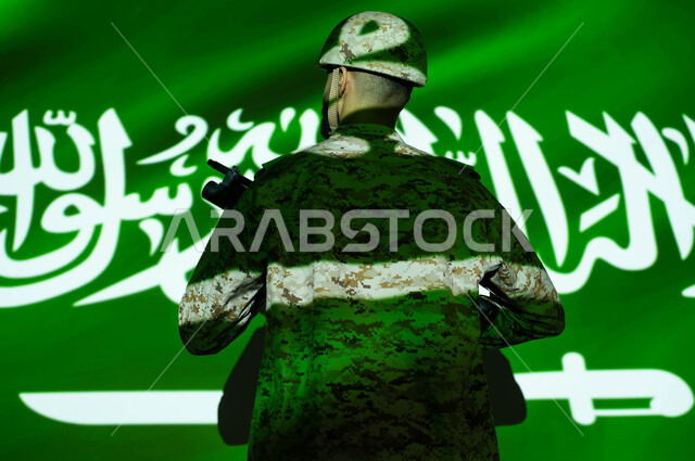 انغماس الجسم في ضوء العلم، تعزيز القوات العسكرية لزيادة الأمن والاستقرار في السعودية، إحياء ذكرى اليوم الوطني السعودي 23 سبتمبر، الاحتفال بالعيد الوطني، يوم العلم 11 مارس، صورة من الخلف لجندي عربي خليجي يرتدي الزي العسكري والخوذة يحمل السلاح.