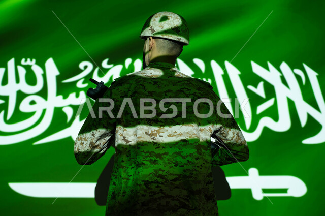 انغماس الجسم في ضوء العلم، تعزيز القوات العسكرية لزيادة الأمن والاستقرار في السعودية، إحياء ذكرى اليوم الوطني السعودي 23 سبتمبر، الاحتفال بالعيد الوطني، يوم العلم 11 مارس، صورة من الخلف لجندي عربي خليجي يرتدي الزي العسكري والخوذة يحمل السلاح.