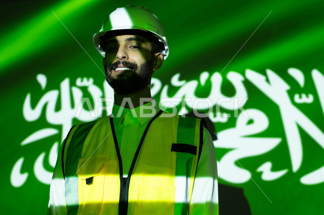 تنمية قطاع الإعمار والتطور الهندسي بسواعد أبناء الوطن, الحب والولاء للبلاد، صورة مقربة لمهندس عربي خليجي سعودي مبتسم يرتدي السترة وخوذة الحماية، انغماس الجسم بضوء العلم، يوم العلم 11 مارس، الإحتفال بذكرى اليوم الوطني السعودي 23 سبتمبر والعيد الوطني