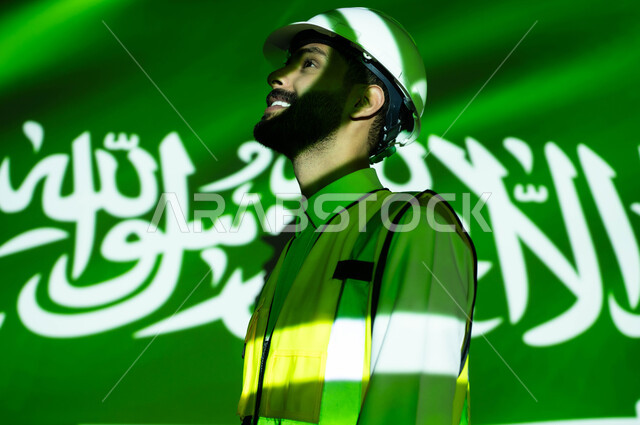 النمو والتطور العمراني للمباني والمنشآت بسواعد أبناء الوطن, صورة مقربة لمهندس عربي خليجي سعودي مبتسم يرتدي السترة وخوذة الحماية، انغماس الجسم بضوء العلم، يوم العلم 11 مارس، الإحتفال بذكرى اليوم الوطني السعودي 23 سبتمبر والعيد الوطني 