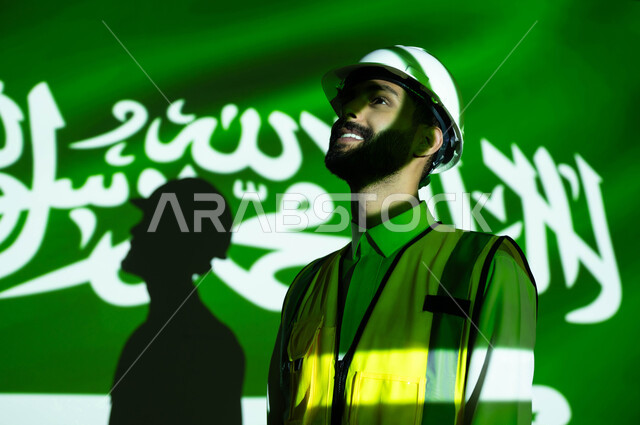 النمو والتطور العمراني للمباني والمنشآت بسواعد أبناء الوطن, صورة مقربة لمهندس عربي خليجي سعودي مبتسم يرتدي السترة وخوذة الحماية، انغماس الجسم بضوء العلم، يوم العلم 11 مارس، الإحتفال بذكرى اليوم الوطني السعودي 23 سبتمبر والعيد الوطني 