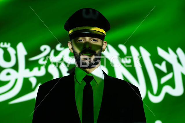 مهنة الملاحة الجوية والطيران في السعودية، الاحتفال بيوم العلم 11 مارس و العيد الوطني، الفخر والاعتزاز بالهوية الوطنية، احياء ذكرى اليوم الوطني السعودي في 23 سبتمبر، صورة مقربة لانعكاس ضوء العلم على وجه كابتن طائرة عربي خليجي سعودي مبتسم يرتدي الزي الخاص بالعمل