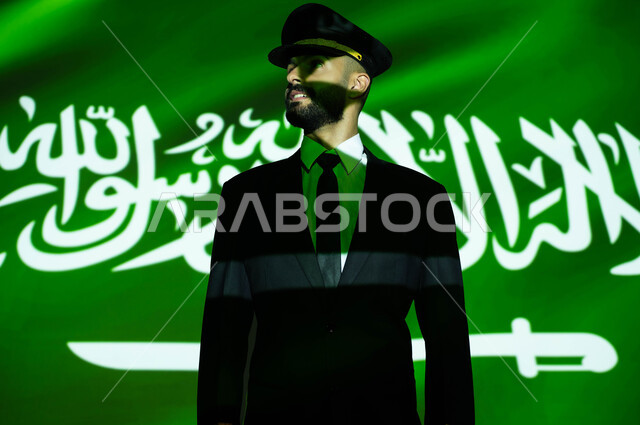 مهنة الملاحة الجوية والطيران في السعودية، الاحتفال بيوم العلم 11 مارس و العيد الوطني، تعزيز الروح الوطنية ،احياء ذكرى اليوم الوطني السعودي في 23 سبتمبر، صورة مقربة لانعكاس ضوء العلم على وجه كابتن طائرة عربي خليجي سعودي مبتسم يرتدي الزي الخاص بالعمل