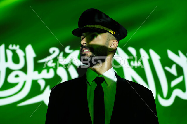 الاحتفال بيوم العلم 11 مارس و العيد الوطني، مهنة الملاحة الجوية والطيران في السعودية، الاخلاص والولاء للبلاد، احياء ذكرى اليوم الوطني السعودي في 23 سبتمبر، صورة مقربة لانعكاس ضوء العلم على وجه كابتن طائرة عربي خليجي سعودي مبتسم يرتدي الزي الخاص بالعمل