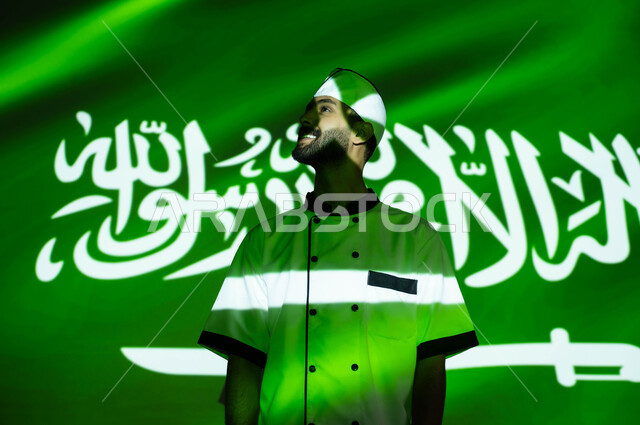 نظرات الفخر والاعتزاز بالبلاد، تطور ونهضة البلاد بالمهن السعوديه المختلفه، الإحتفال في العيد الوطني و يوم العلم 11 مارس، اليوم الوطني السعودي 8 ربيع الأول، صورة مقربة لطباخ عربي خليجي سعودي مبتسم يرتدي زي الشيف، اندماج الوجه بضوء العلم، الحب والاخلاص للوطن
