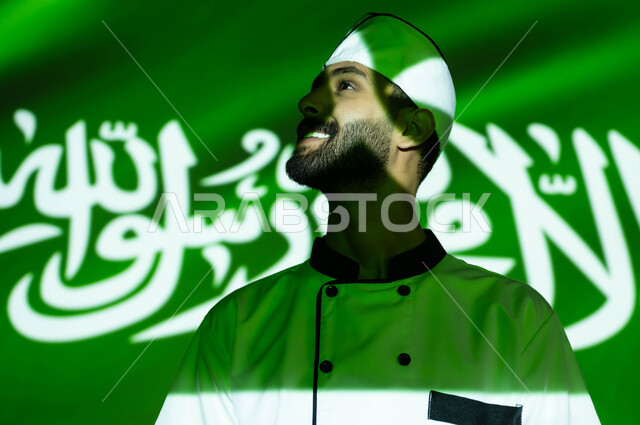 نظرات الفخر والاعتزاز بالبلاد، تطور ونهضة البلاد بالمهن السعوديه المختلفه، الإحتفال في العيد الوطني و يوم العلم 11 مارس، اليوم الوطني السعودي 8 ربيع الأول، صورة مقربة لطباخ عربي خليجي سعودي مبتسم يرتدي زي الشيف، اندماج الوجه بضوء العلم، الحب والاخلاص للوطن
