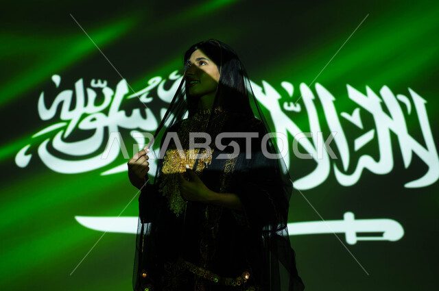 Pride and pride in the achievements of the nation and its culture, Flag Day, March 11, celebration of the anniversary of the Saudi National Day, September 23, a smiling Saudi Gulf Arab woman wearing the sheila and the dress of the central region on National Day, national celebrations and events, the reflection of the light of the flag on the face, Founding Day 1727 AD