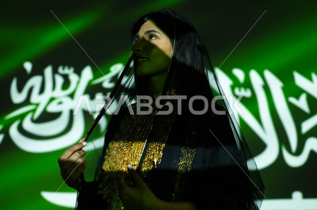 الفخر بإنجازات الوطن وثقافته، يوم العلم 11 مارس، الاحتفال بذكرى اليوم الوطني السعودي 23 سبتمبر، صورة مقربة لإمرأة عربية خليجية سعودية مبتسمة ترتدي الشيلة وثوب المنطقة الوسطى في العيد الوطني، احتفالات ومناسبات وطنية، انعكاس ضوء العلم على الوجه، يوم التأسيس 1727 م