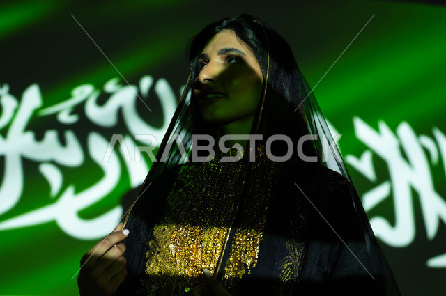 Pride in the achievements of the nation and its culture, Flag Day, March 11, celebration of the anniversary of the Saudi National Day, September 23, a close-up of a smiling Saudi Gulf Arab woman wearing the sheila and the dress of the central region on National Day, national celebrations and events, the reflection of the light of the flag on the face, Founding Day 1727 AD