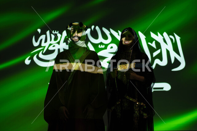 استذكار تراث وتاريخ الاجداد بارتداء الأزياء الشعبية الوطنية، ذكرى اليوم الوطني السعودي في 23 سبتمبر، انعكاس ضوء العلم على جسد رجل وامرأة عربيان خليجيان سعوديان مبتسمان يرتديان زي المنطقة الوسطى التقليدي في العيد الوطني، يوم العلم 11 مارس، يوم التأسيس 1727 م
