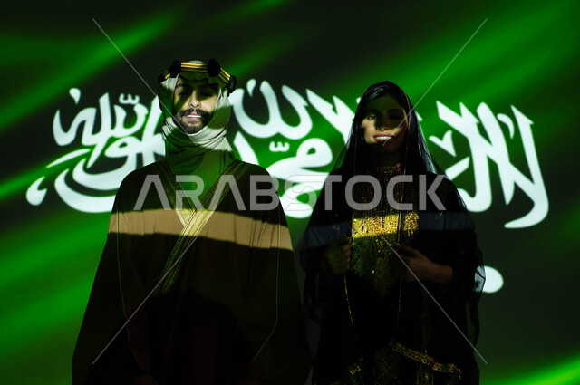 استذكار تراث وتاريخ الاجداد بارتداء الأزياء الشعبية الوطنية، ذكرى اليوم الوطني السعودي في 23 سبتمبر، انعكاس ضوء العلم على جسد رجل وامرأة عربيان خليجيان سعوديان مبتسمان يرتديان زي المنطقة الوسطى التقليدي في العيد الوطني، يوم العلم 11 مارس، يوم التأسيس 1727 م