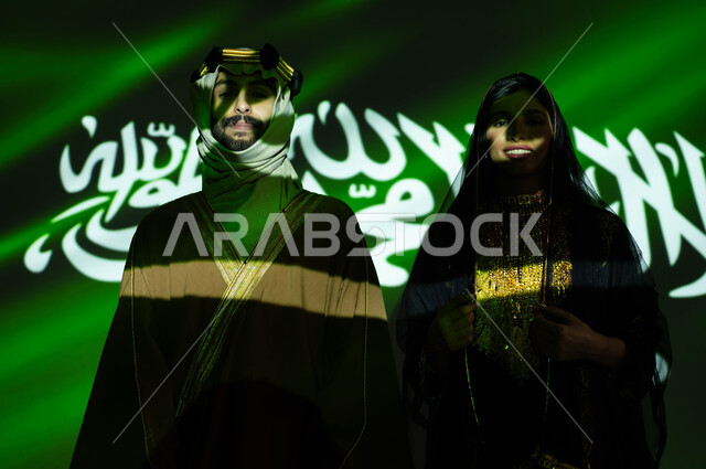 استذكار تاريخ وتراث الاجداد بارتداء الأزياء الشعبية الوطنية، ذكرى اليوم الوطني السعودي في 23 سبتمبر، صورة مقربة لانعكاس ضوء العلم على جسد رجل وامرأة عربيان خليجيان سعوديان مبتسمان يرتديان زي المنطقة الوسطى في العيد الوطني، يوم العلم 11 مارس، يوم التأسيس 1727 م