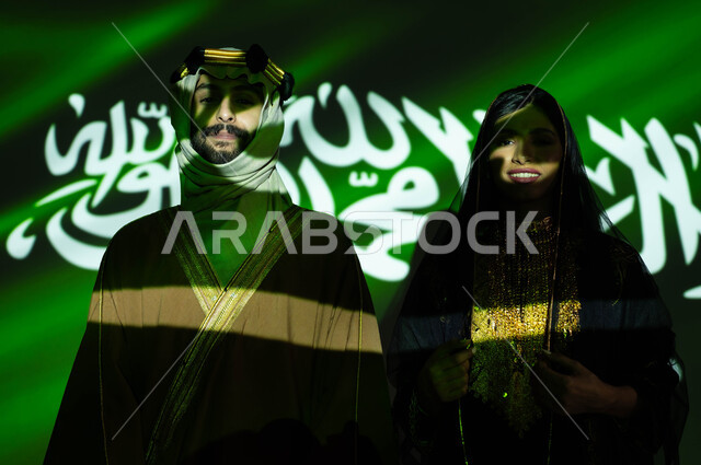 استذكار تاريخ وتراث الاجداد بارتداء الأزياء الشعبية الوطنية، ذكرى اليوم الوطني السعودي في 23 سبتمبر، صورة مقربة لانعكاس ضوء العلم على جسد رجل وامرأة عربيان خليجيان سعوديان مبتسمان يرتديان زي المنطقة الوسطى في العيد الوطني، يوم العلم 11 مارس، يوم التأسيس 1727 م