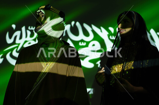 صورة مقربة لانعكاس ضوء العلم على جسد رجل وامرأة عربيان خليجيان سعوديان مبتسمان يرتديان زي المنطقة الوسطى في العيد الوطني، يوم العلم 11 مارس، الحفاظ على تراث الاجداد بارتداء الأزياء الشعبية الوطنية، ذكرى اليوم الوطني السعودي في 23 سبتمبر، يوم التأسيس 1727 م