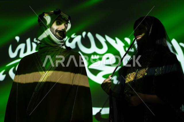 الحفاظ على تراث وهوية الاجداد بارتداء الأزياء الشعبية الوطنية، انعكاس ضوء العلم على جسد رجل وامرأة عربيان خليجيان سعوديان مبتسمان يرتديان زي المنطقة الوسطى في العيد الوطني، ذكرى اليوم الوطني السعودي في 23 سبتمبر، الاحتفال بيوم العلم 11 مارس، يوم التأسيس 1727 م