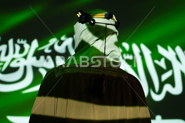 انغماس الجسد في ضوء العلم، إحياء ذكرى اليوم الوطني السعودي 8 ربيع الاول، تأسيس الدولة السعودية 1727 م ،اظهار التفاني للبلاد، يوم العلم 11 مارس، صورة مقربة من الخلف لرجل عربي خليجي سعودي يرتدي البشت والغترة في العيد الوطني، الزي الشعبي التقليدي للمنطقة الجنوبية