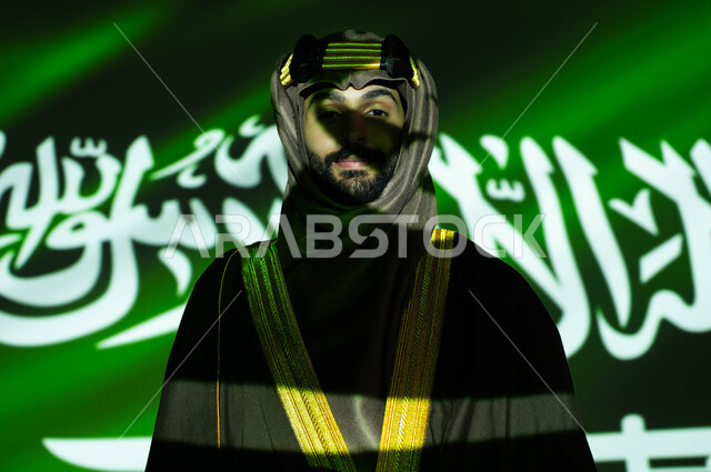 الفخر والاعتزاز بإنجازات الوطن وثقافته، صورة مقربة لانعكاس ضوء العلم على وجه رجل عربي خليجي سعودي مبتسم يرتدي البشت والغترة في العيد الوطني، الزي الشعبي التقليدي للمنطقة الجنوبية، احياء ذكرى اليوم الوطني السعودي 23 سبتمبر، يوم العلم 11 مارس، يوم التأسيس 1727 م