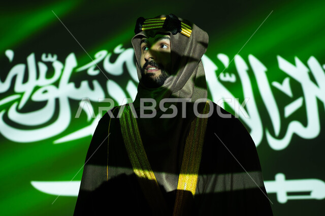 صورة مقربة لانعكاس ضوء العلم على وجه رجل عربي خليجي سعودي يرتدي البشت والغترة في العيد الوطني، الفخر والاعتزاز بإنجازات الوطن وثقافته، الزي الشعبي التقليدي للمنطقة الجنوبية، احياء ذكرى اليوم الوطني السعودي 23 سبتمبر، يوم العلم 11 مارس، يوم التأسيس 1727 م