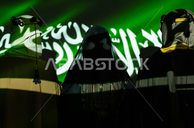 انعكاس ضوء العلم على الجسم، صورة مقربة من الخلف لمواطنين عرب خليجيين سعوديين يرتدون الزي التقليدي الشعبي، يوم العلم 11 مارس، مشاعر الحب والولاء والانتماء للمملكة،  الإحتفال بالمناسبات وذكرى اليوم الوطني السعودي 23 سبتمبر، تأسيس الدولة السعودية الاولى 1727 م