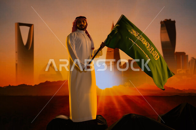 النمو والتطور العمراني في السعودية، رجل عربي خليجي سعودي يرتدي الثوب الأبيض والشماغ ويحمل علم السعودية بفخر، ذكرى اليوم الوطني لتوحيد المملكة 23 سبتمبر، يوم العلم 11 مارس، العيد الوطني السعودي، خلفية الأبراج وناطحات السحاب وقت غروب الشمس
