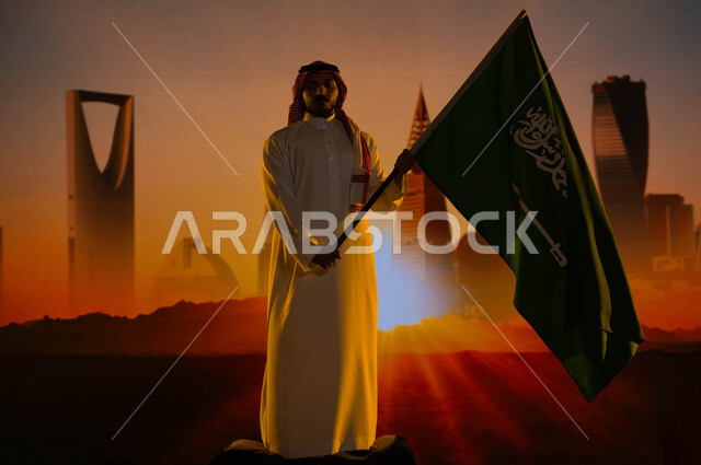 رؤية السعودية المستقبلية 2030، رجل عربي خليجي سعودي يرتدي الثوب الأبيض والشماغ ويحمل علم السعودية، ذكرى اليوم الوطني لتوحيد المملكة 23 سبتمبر، يوم العلم السعودي 11 مارس، العيد الوطني السعودي، خلفية أبراج وناطحات سحاب مدينة الرياض وقت غروب الشمس