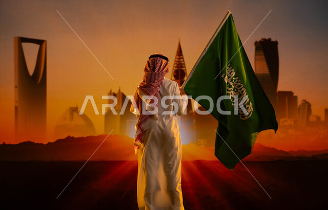 يوم العلم السعودي 11 مارس، اليوم الوطني لتوحيد السعودية، صورة من الخلف لولد عربي خليجي سعودي يرتدي الثوب الأبيض والشماغ ويحمل علم السعودية ويقف امام أبراج وناطحات سحاب الرياض وقت شروب الشمس، العيد الوطني 8 ربيع الأول، رؤية 2030 المستقبلية
