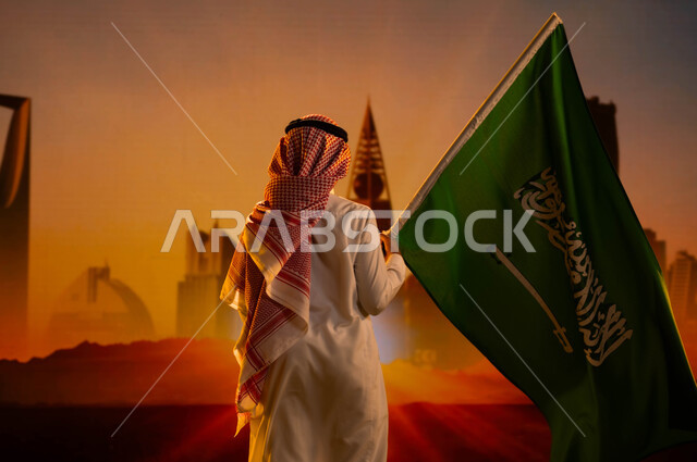 النمو والتطور العمراني في  الرياض، الفخر والاعتزاز بالوطن، اليوم الوطني السعودي 23 سبتمبر، صورة من الخلف لولد عربي خليجي سعودي يرتدي الثوب والشماغ ويحمل علم السعودية، الإحتفال بالعيد الوطني، يوم العلم السعودي 11 مارس، رؤية السعودية المستقبلية 2030