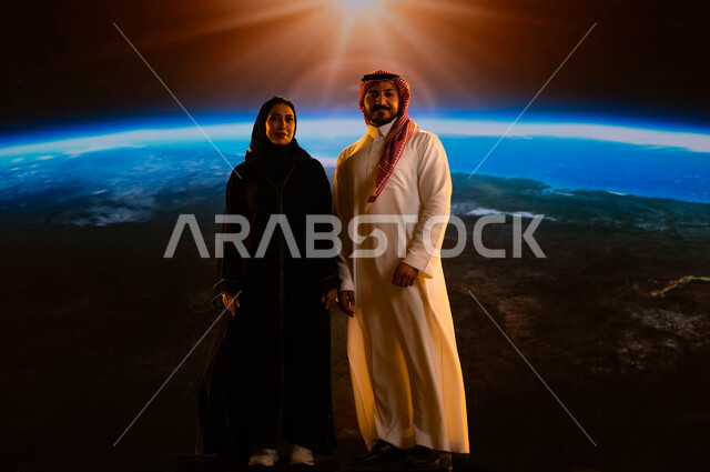 زوجان عربيان خليجيان سعوديان يقفان بثقة وينظران إلى الكاميرا، نمو وتطور قطاع الفضاء في المملكة، رحلة السعودية إلى الفضاء الخارجي، مفهوم الترابط الأسري، إيماءات السرور والسعادة، صورة الجسم بالطول الكامل، خلفية شروق الشمس على الكرة الأرضية