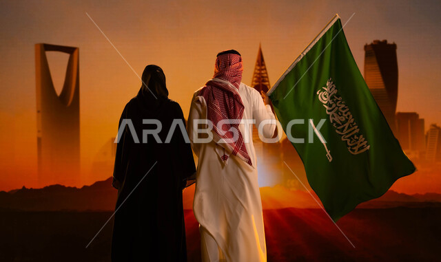 التطور العمراني لناطحات السحاب في السعودية، صورة من الخلف لرجل عربي خليجي سعودي يقف بجانب زوجته ويحمل علم المملكة، العيد الوطني لتوحيد السعودية، اليوم الوطني السعودي 23 سبتمبر، يوم العلم 11 مارس، رؤية 2030 المستقبلية، غروب الشمس على أبراج الرياض
