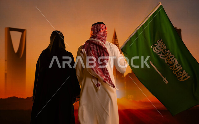رؤية السعودية 2030، التقدم والتطور العمراني، صورة من الخلف لرجل عربي خليجي سعودي يقف بجانب زوجته ويحمل علم المملكة، الوقوف اما الأبراج وناطحات سحاب الرياض وقت غروب الشمس، اليوم الوطني السعودي، العيد الوطني لتوحيد السعودية 8 ربيع الأول، يوم العلم 11 مارس، 