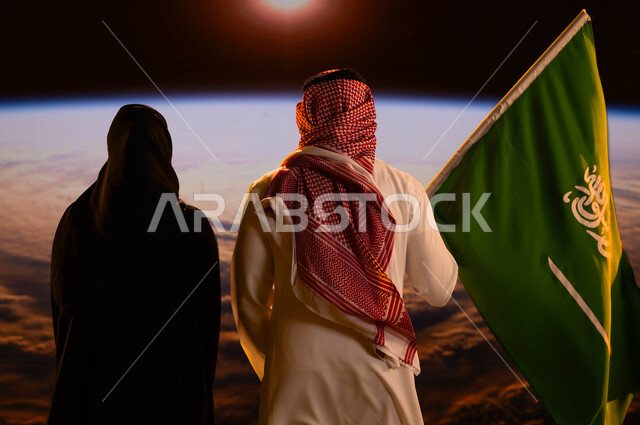 تطور ونمو قطاع الفضاء، صورة من الخلف لرجل عربي خليجي سعودي يقف بجانب زوجته ويحمل علم السعودية، الهوية الوطنية التاريخية للبلاد، اليوم الوطني لتوحيد المملكة 8 ربيع الأول، الإحتفال بالعيد الوطني السعودي، يوم العلم السعودي 11 مارس، شروق الشمس على كوكب الأرض