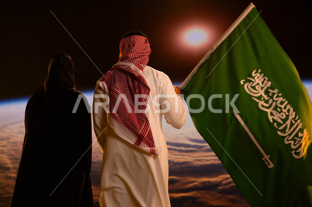 اليوم الوطني لتوحيد المملكة 23 سبتمبر، صورة من الخلف لرجل عربي خليجي سعودي يقف بجانب زوجته ويحمل علم السعودية، حب الوطن والحفاظ على هوية البلاد، ذكرى العيد الوطني السعودي، يوم العلم 11 مارس، تطور ونمو قطاع الفضاء، خلفية شروق الشمس على الكرة الأرضية