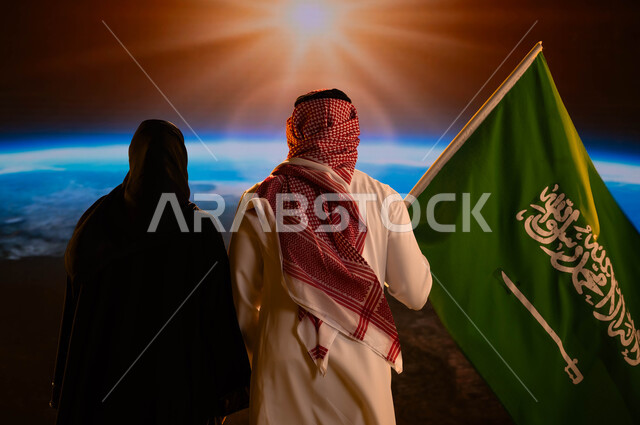 رحلة السعودية إلى الفضاء الخارجي، صورة من الخلف لزوجان عربيان خليجيان سعوديان، رجل يرتدي الزي السعودي التقليدي ويحمل علم المملكة إحتفالا باليوم الوطني لتوحيد السعودية، العيد الوطني 8 ربيع الأول، يوم العلم السعودي 11 مارس، شروق الشمس على كوكب الأرض