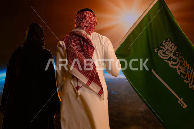 العيد الوطني السعودي 23 سبتمبر، يوم العلم 11 مارس، صورة من الخلف لزوجان عربيان خليجيان سعوديان، رجل يرتدي الزي السعودي التقليدي ويحمل علم المملكة إحتفالا بذكرى اليوم الوطني، مفهوم  حب الوطن، شروق الشمس على الكرة الأرضية