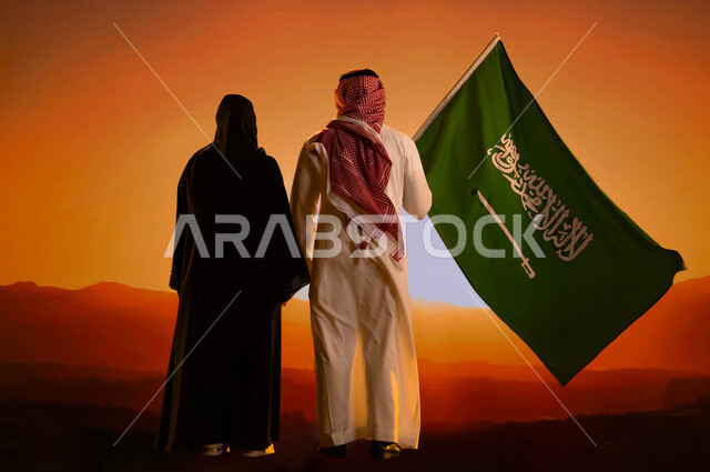 Preserving national identity, a photo from the back of a Saudi Gulf Arab man standing next to his wife and holding the flag of the Kingdom, Flag Day, March 11, Saudi National Day, National Day for the Unification of Saudi Arabia, September 23, full-length body photo, mountain peaks and heights at sunset