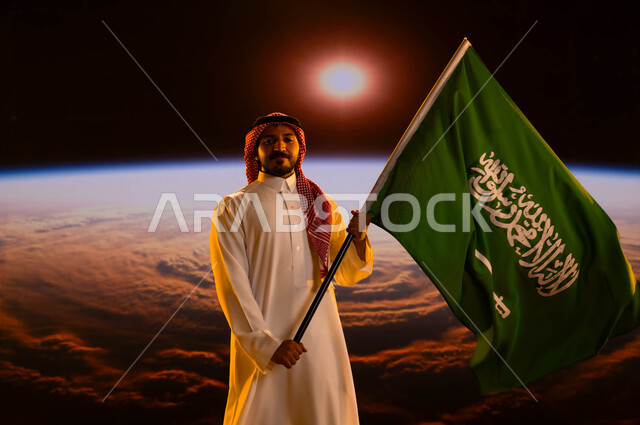 التعبير عن الحب والولاء للوطن، رجل عربي خليجي سعودي يرتدي الثوب الابيض والشماغ ينظر إلى الكاميرا ويحمل علم السعودية، اليوم الوطني لتوحيد السعودية، الإحتفال بيوم العلم 11 مارس، ذكرى العيد الوطني 23 سبتمبر، خلفية شروق الشمس على الأرض