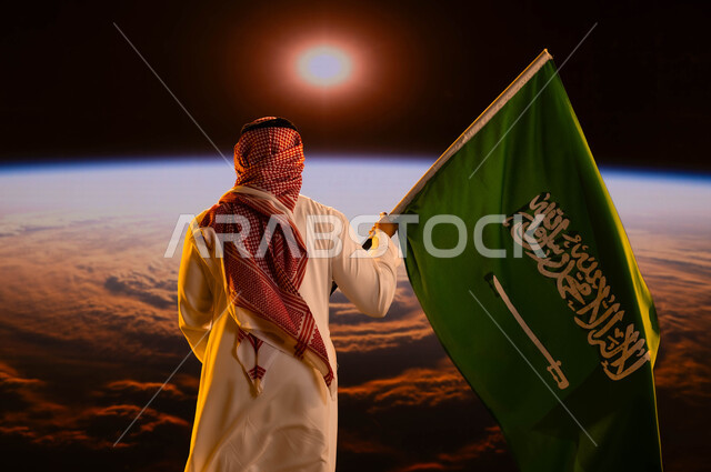 يوم العلم السعودي11 مارس، صورة من الخلف لرجل عربي خليجي سعودي يرتدي الشماغ والثوب الأبيض ويحمل علم المملكة، النظر لكوكب الأرض في الفضاء الخارجي، التعبير عن الحب والولاء للوطن، اليوم الوطني السعودي، العيد الوطني 8 ربيع الاول، شروق الشمس على الكرة الأرضية
