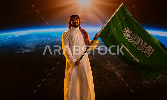ذكرى العيد الوطني 23 سبتمبر، رجل عربي خليجي سعودي يرتدي الشماغ والثوب الابيض ينظر للكاميرا ويحمل علم السعودية بفخر وإعتزاز، التفاني والإنتماء للوطن، الإحتفال باليوم الوطني لتوحيد السعودية، يوم العلم السعودي 11 مارس، خلفية الكرة الأرضية في الفضاء الخارجي