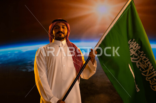 ذكرى اليوم الوطني السعودي، رجل عربي خليجي سعودي يرتدي الزي التقليدي ينظر إلى الكاميرا ويحمل علم السعودية بفخر وإعتزاز، مفهوم  حب الوطن، الإحتفال بيوم العلم 11 مارس، العيد الوطني 23 سبتمبر، رحلة المملكة إلى الفضاء الخارجي، خلفية شروق الشمس