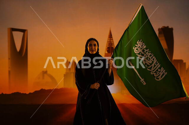 مفهوم الإنشاءات والهندسة المعمارية، امرأة عربية خليجية سعودية محجبة ترتدي العباءة وتحمل علم المملكة، إستذكار التاريخ والهوية الوطنية، يوم العلم 11 مارس، اليوم الوطني لتوحيد السعودية، العيد الوطني السعودي 23 سبتمبر، خلفية الأبراج وناطحات السحاب في الرياض