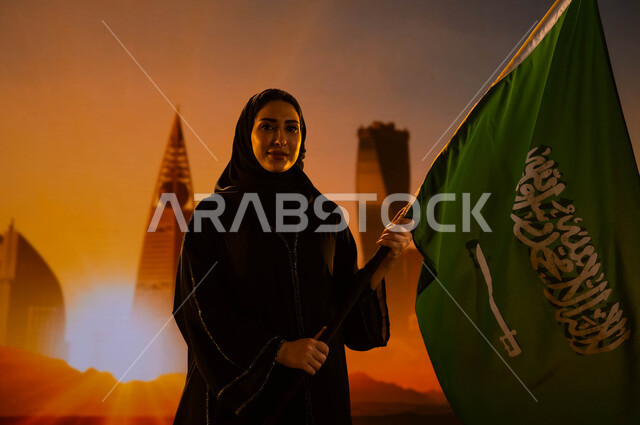 النمو والتطور العمراني في السعودية، امرأة عربية خليجية سعودية محجبة ترتدي العباءة وتحمل علم المملكة، إيماءات الفخر والاعتزاز بالوطن، يوم العلم 11 مارس، اليوم الوطني لتوحيد السعودية، العيد الوطني 23 سبتمبر، خلفية شروق الشمس على أبراج الرياض