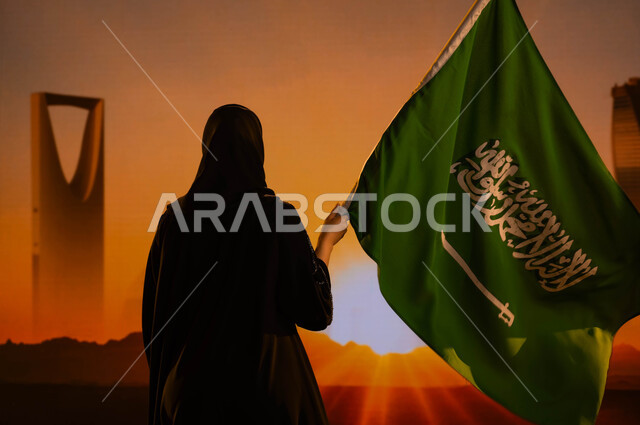 ذكرى اليوم الوطني 8 ربيع الاول، صورة من الخلف لإمرأة عربية خليجية سعودية محجبة ترتدي العباءة وتحمل علم المملكة وتقف امام أبراج وناطحات سحاب الرياض، يوم العلم السعودي 11 مارس، العيد الوطني السعودي، معالم مدينة الرياض وقت شروق الشمس