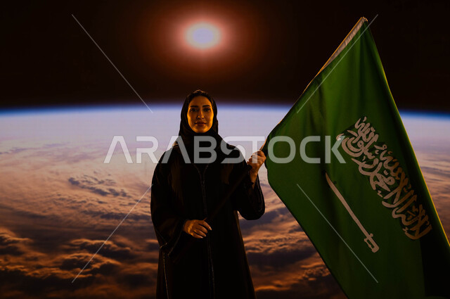 الوقوف بثقة والنظر إلى الكاميرا، امرأة عربية خليجية سعودية مبتسمة ترتدي العباءة والحجاب وتحمل علم السعودية بفخر وإعتزاز، الإحتفال بذكرى اليوم الوطني السعودي، يوم العلم 11 مارس، العيد الوطني السعودي 23 سبتمبر، خلفية شروق الشمس على الكرة الأرضية