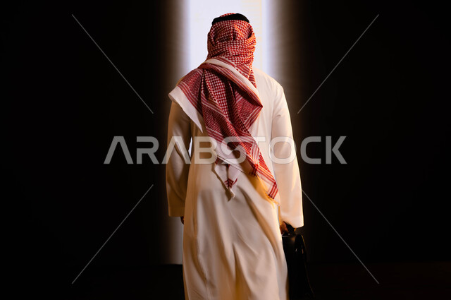 التحرر من القيود والانتقال من الظلام إلى النور، صورة من الخلف لرجل أعمال عربي سعودي خليجي يرتدي الزي التقليدي يحمل بيده حقيبة اللابتوب ويقوم  بالدخول الى خلفية بيضاء، رؤية سعودية 2030،  فتح آفاق جديدة للسعوديين، مساهمة رجال الاعمال في تقدم وتطور البلاد