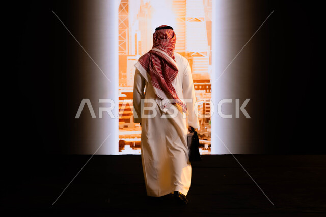 Pride in the achievements achieved thanks to the unification of the country, implementing urban development projects in Saudi Arabia, Saudi Vision 2030, towers and urban achievements in Riyadh, a picture from behind of a Saudi Gulf Arab man wearing a shemagh and traditional thobe, holding a handbag and standing in front of the Riyadh towers.