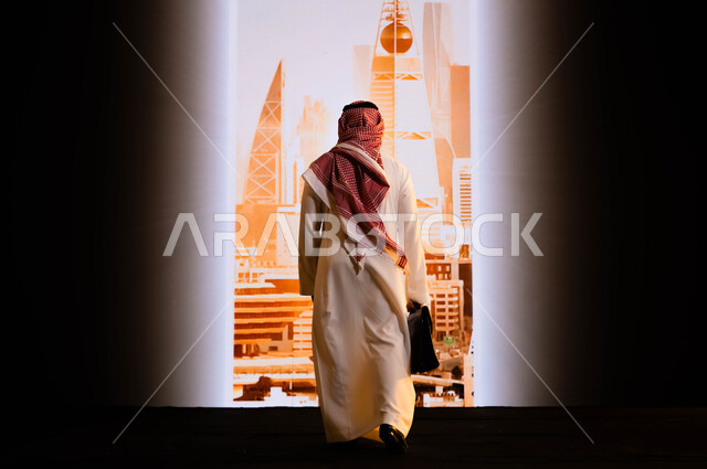 الأبراج و الإنجازات العمرانية في الرياض , تنفيذ مشاريع تنموية عمرانية في السعودية, رؤية سعودية 2030, الافتخار بالإنجازات التي تحققت بفضل توحيد البلاد, صورة من الخلف لرجل عربي سعودي خليجي يرتدي الشماغ والثوب التقليدي و يمسك حقيبة يد ويقف امام أبراج الرياض