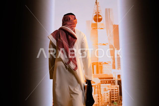 Saudi Vision 2030, implementing urban development projects in Saudi Arabia, towers and urban achievements in Riyadh, pride in the achievements achieved thanks to the unification of the country, a picture from behind of a Saudi Gulf Arab man wearing a shemagh and traditional thobe, holding a handbag and standing in front of the Riyadh towers